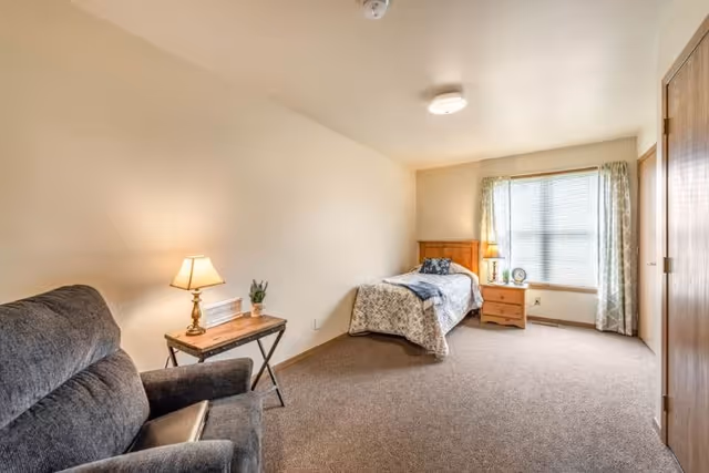 A simple, cozy bedroom in a senior living facility with a single bed covered in a floral quilt, a wooden nightstand with a lamp and decorative items, a large window with patterned curtains, a comfortable gray armchair, and a small wooden side table with a lamp and a plant.
