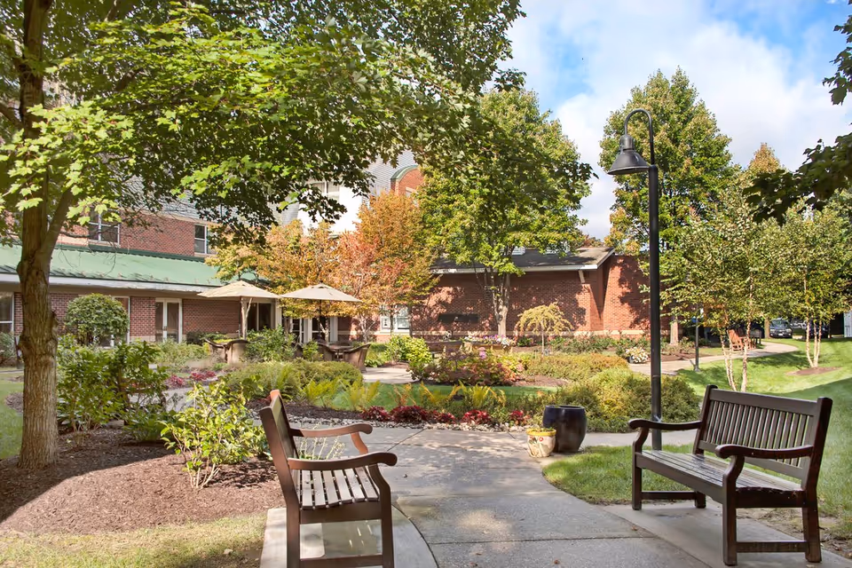 Outdoor garden area at Brighton Gardens of Columbia featuring a paved walkway, wooden benches, a variety of trees and shrubs, patio tables with umbrellas, and a brick building in the background under a partly cloudy sky.