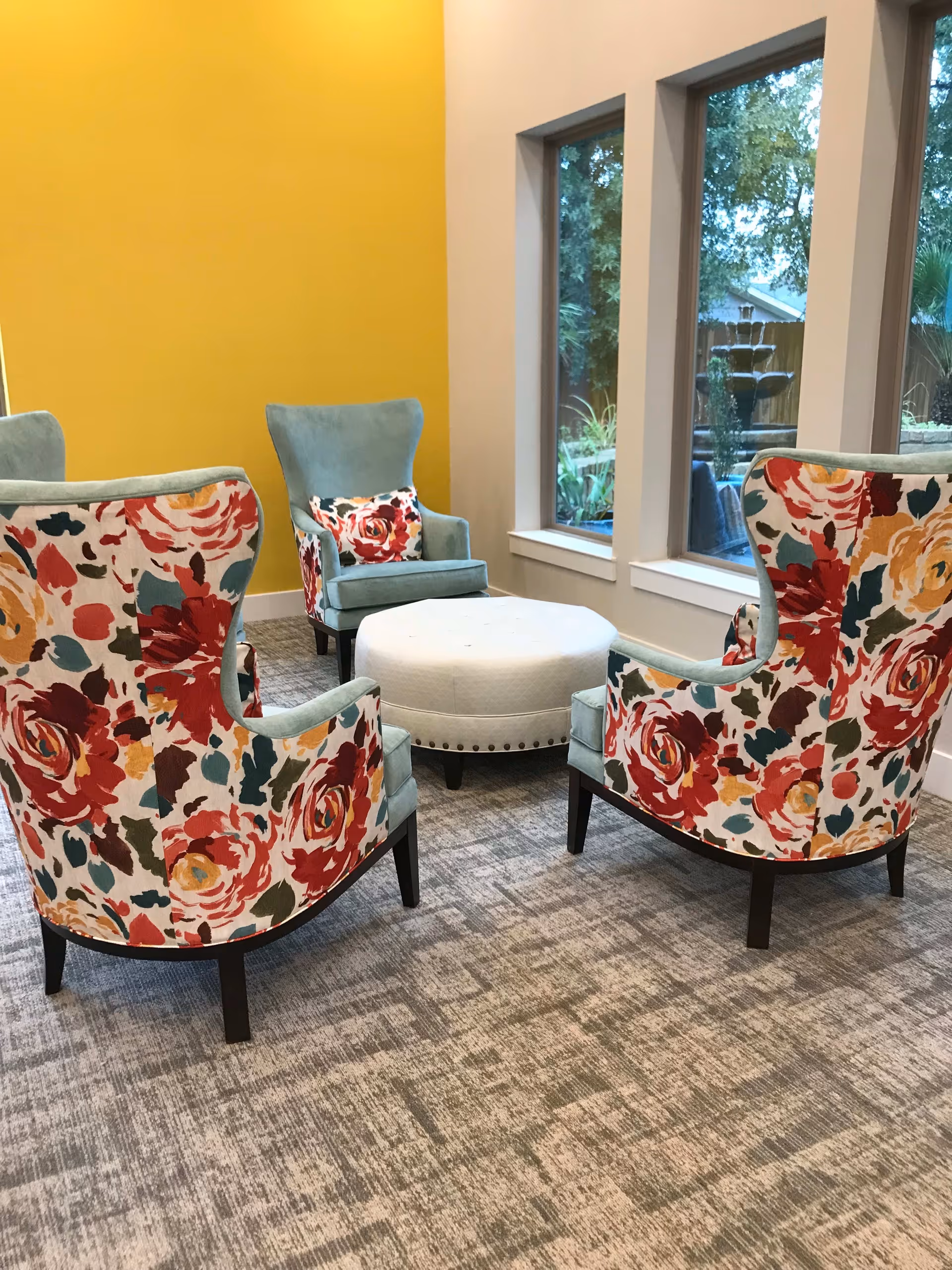 A cozy seating area with four armchairs arranged around a round white ottoman. Two of the chairs have a floral pattern with red, yellow, and teal colors, while the other two are solid teal with matching floral pillows. The room has a bright yellow accent wall and three large windows showing an outdoor garden with a water fountain.