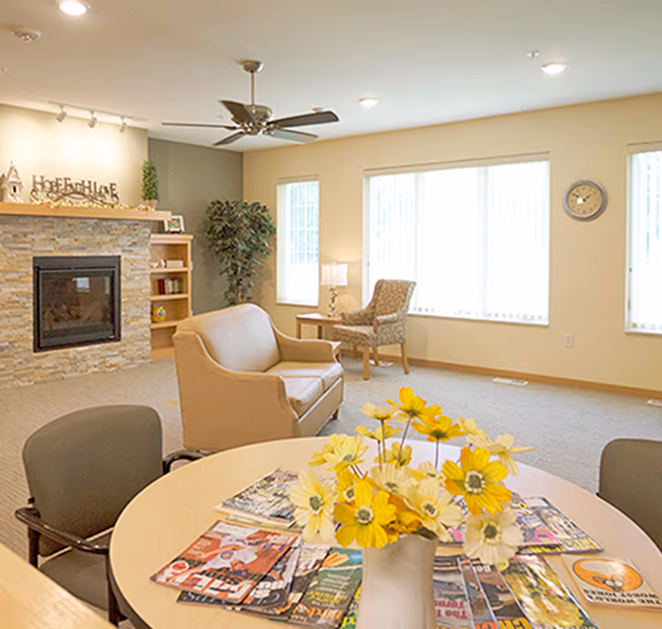 Bright communal living room with seating, a fireplace, a table of yellow flowers and magazines, and large windows.