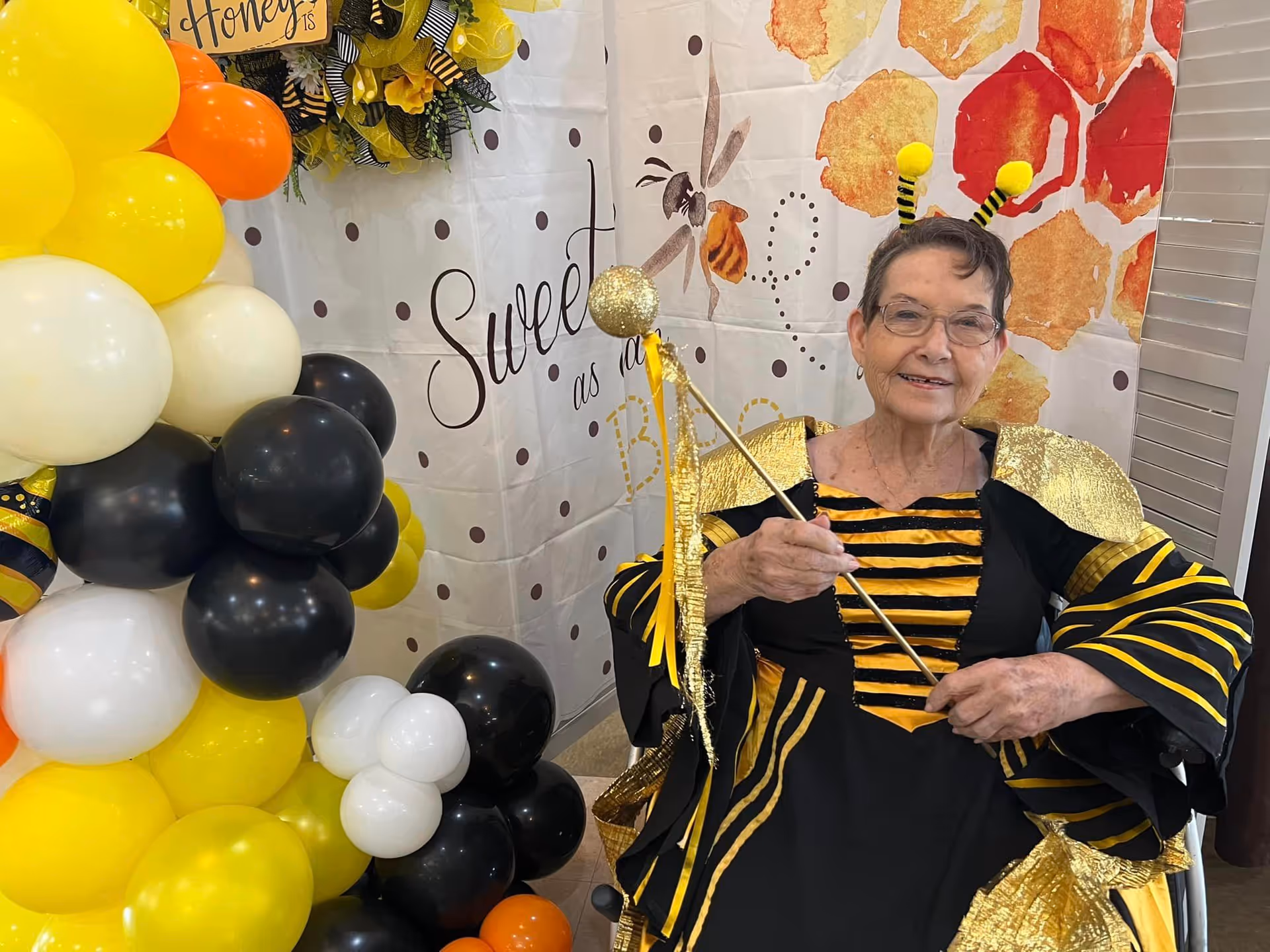 An elderly woman dressed in a black and yellow bee costume with antennae headband and golden wings, sitting and smiling in front of a backdrop decorated with honeycomb patterns and the words 'Sweet as a Bee'. There is a colorful balloon arrangement in yellow, black, white, and orange colors to her left.