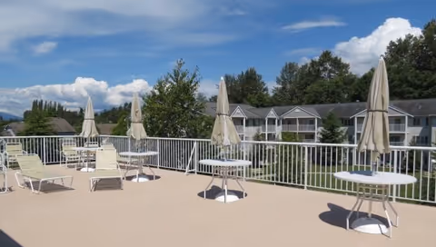 Outdoor patio area with several round tables and closed umbrellas, along with lounge chairs. The patio is surrounded by a white railing and overlooks a residential building with balconies, trees, and a partly cloudy sky.