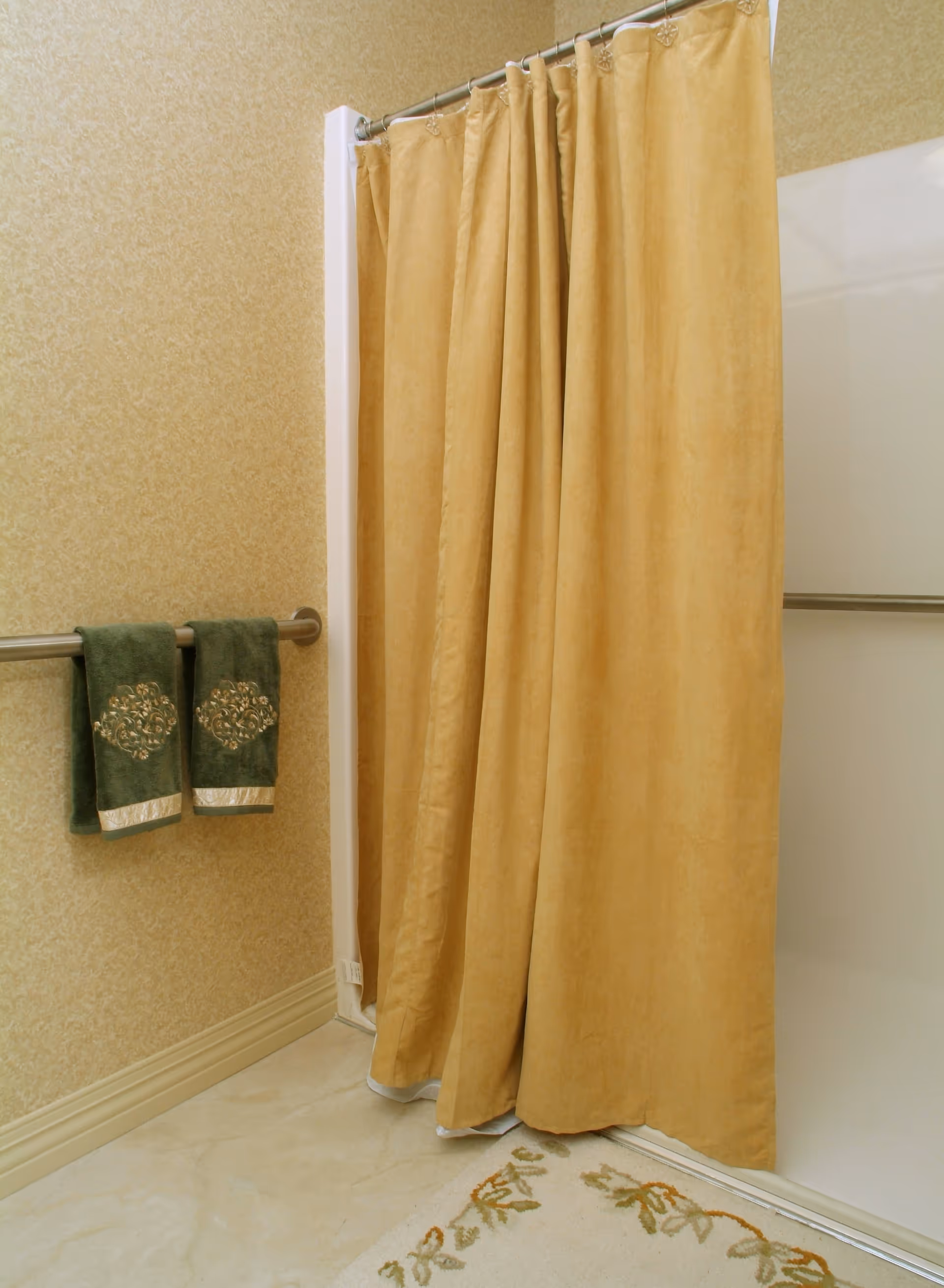 A bathroom shower area with a beige shower curtain partially closed. There are two green towels with decorative patterns hanging on a metal towel bar mounted on a beige patterned wall. A floral-patterned bath mat is on the floor in front of the shower.
