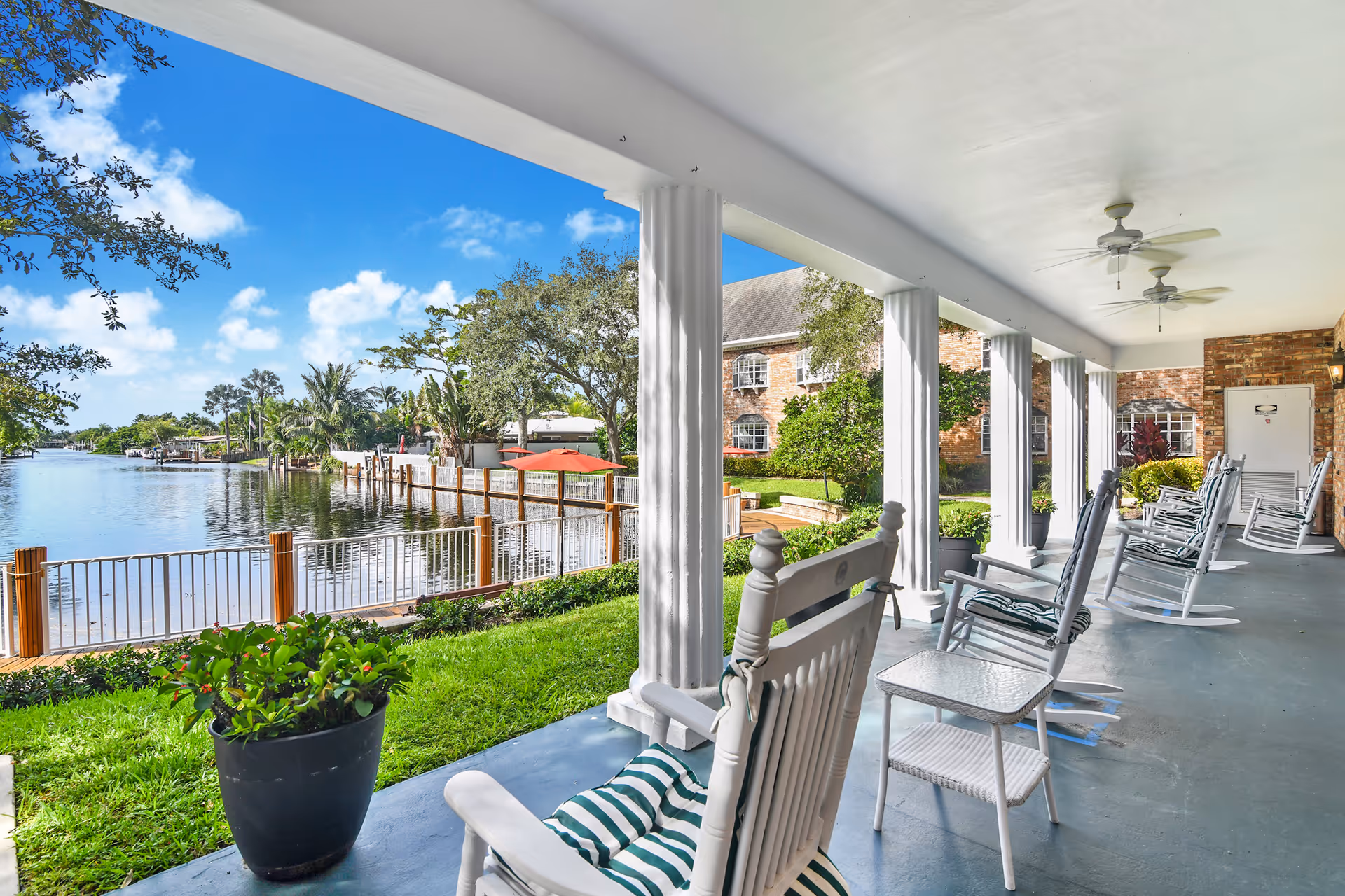 Covered porch with white columns and several rocking chairs facing a calm canal and dock.