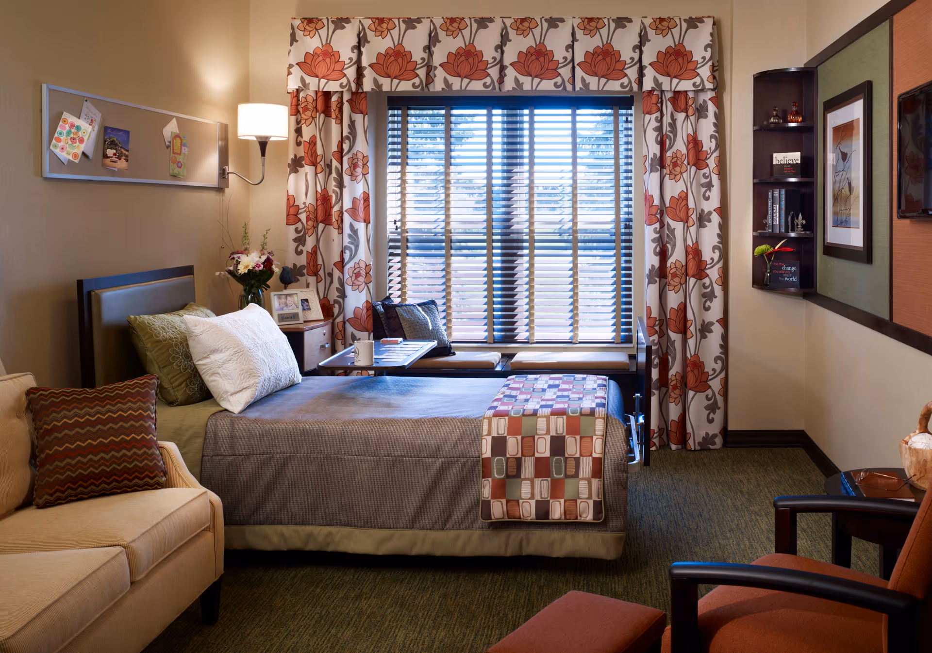 A cozy bedroom in a senior living facility featuring a single bed with pillows and a patterned throw, a beige sofa with a decorative pillow, a window with floral curtains and blinds, a small table with a cup and books, a wall-mounted lamp, and a corner shelf with decorative items and framed artwork on the walls.