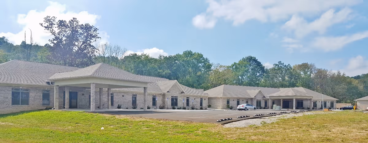 Front exterior of a single-story brick senior living facility with a covered entrance, parking area, and surrounding trees.