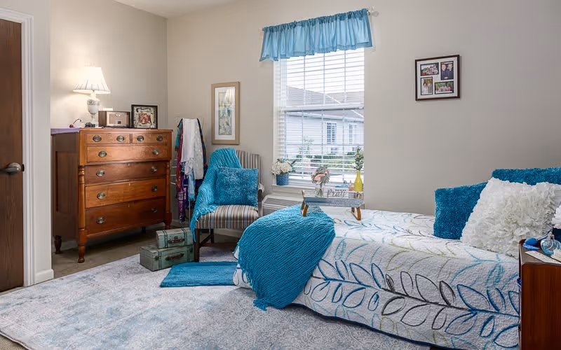 A cozy bedroom with a single bed covered in a white and blue leaf-patterned quilt and blue throw blanket. There are blue and white pillows on the bed. Next to the bed is a wooden nightstand with a lamp and some items. A window with white blinds and a blue valance lets in natural light. A striped armchair with a blue throw blanket and pillow sits near the window. A wooden dresser with a lamp, framed photos, and decorative items is against the wall. The room has a light-colored carpet and neutral walls with framed pictures.