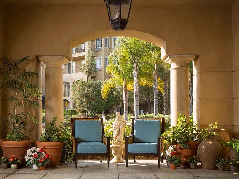Covered courtyard seating area with two blue cushioned chairs flanking a small statue, surrounded by potted plants and columns overlooking palm trees.