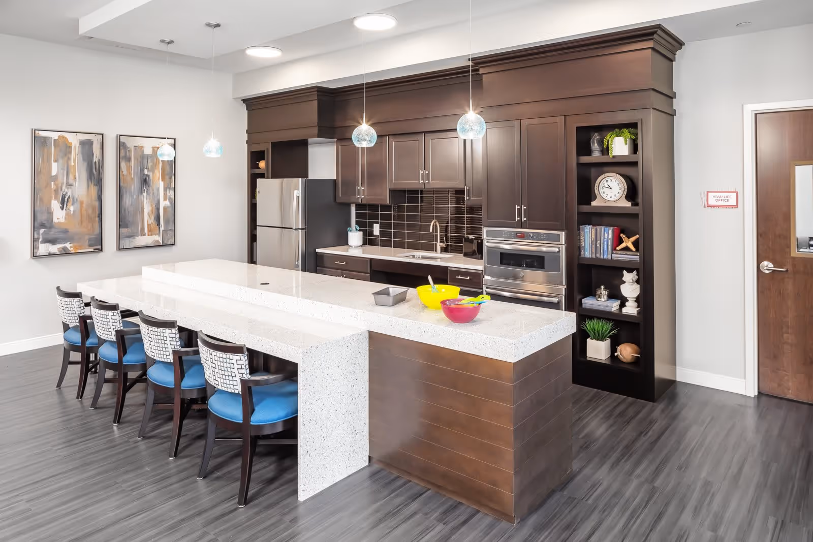 Modern kitchen area with dark wood cabinets, stainless steel refrigerator and oven, a white quartz countertop island with four chairs featuring blue cushions. The kitchen has pendant lights hanging above the island, two abstract paintings on the wall, and a built-in bookshelf with decorative items and books.