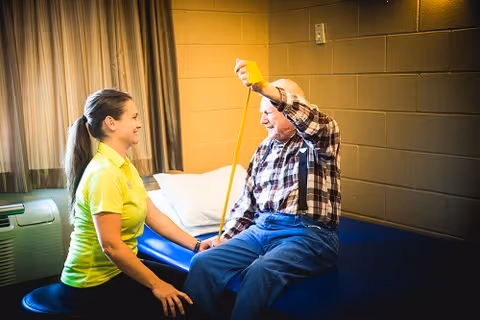A caregiver in a yellow shirt assists an elderly man sitting on a blue examination table as he measures his arm with a yellow measuring tape in a room with beige walls and a window with curtains.