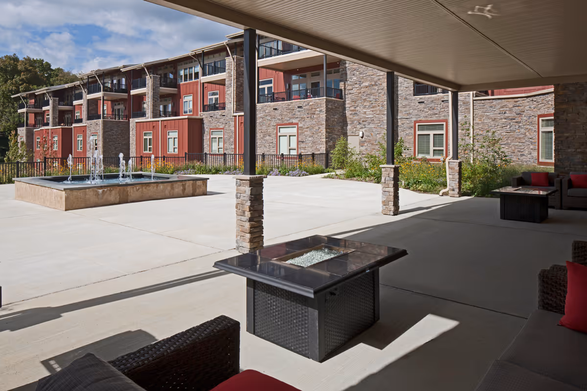Covered patio with seating and a fire table overlooking a courtyard fountain and a multi-story stone-and-red building.