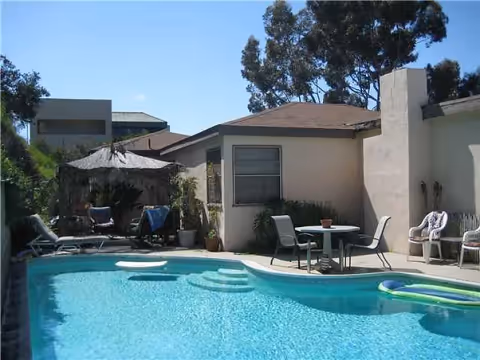 Outdoor swimming pool area with clear blue water, surrounded by a patio with chairs, a table, and a shaded seating area. The pool has steps leading into the water and a pool float. Trees and a building are visible in the background.