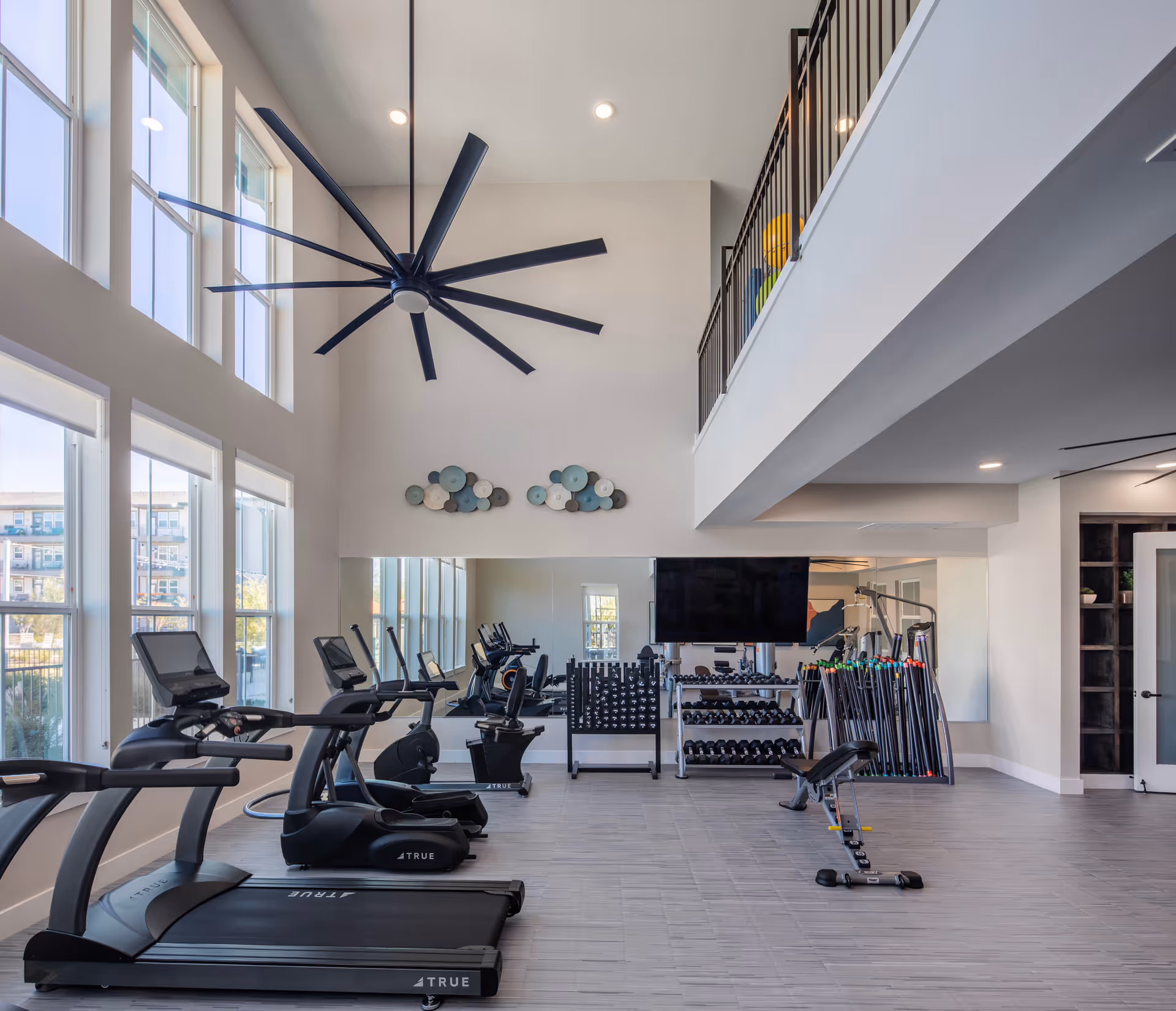 A modern fitness room with large windows letting in natural light, featuring treadmills, exercise bikes, a weight bench, dumbbells, and other workout equipment. The room has high ceilings with a large black ceiling fan and a mezzanine with a railing above.