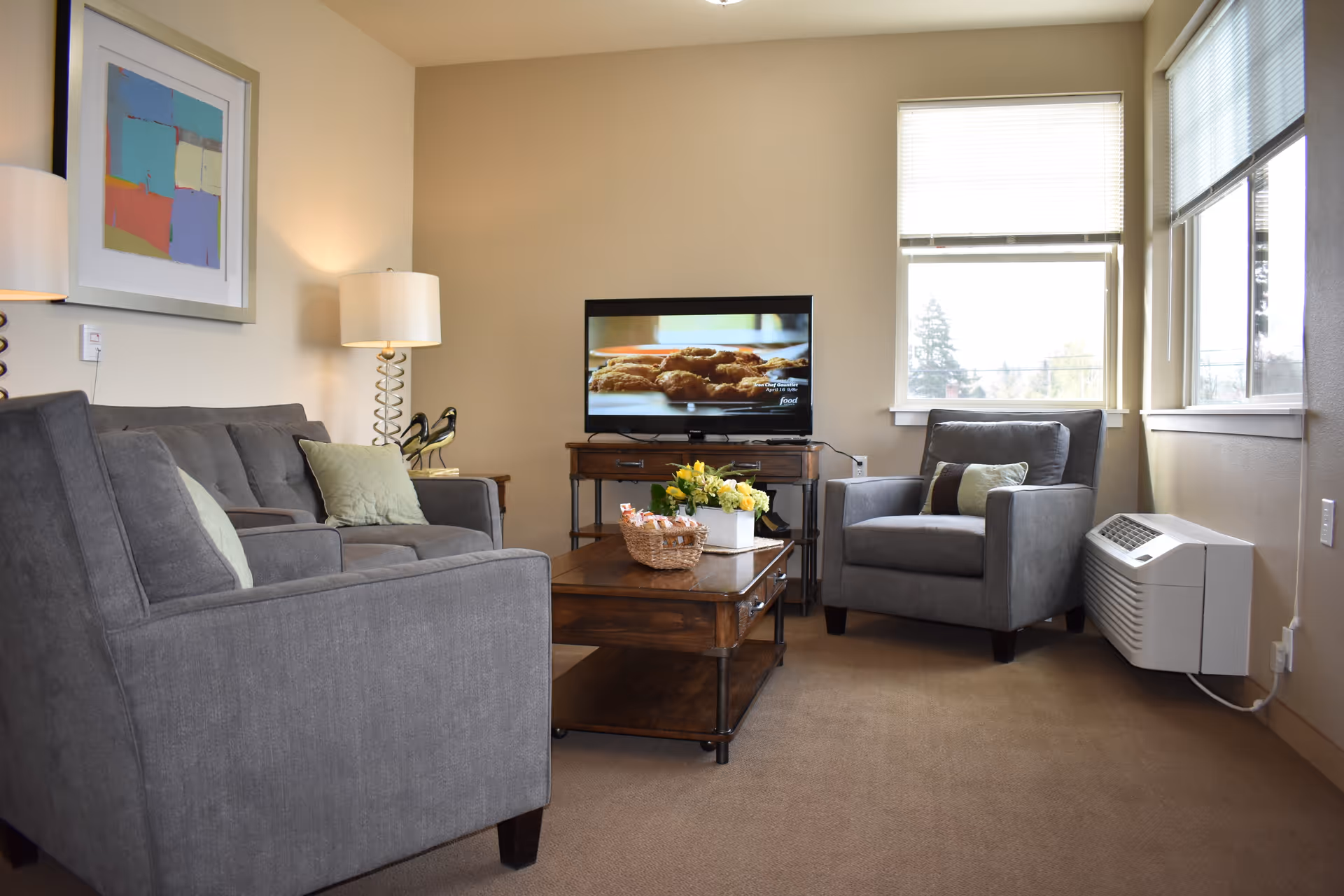 A cozy living room with a gray sofa and armchair, both with light green and brown cushions. A wooden coffee table with a basket and a flower pot sits in the center. A flat-screen TV is on a wooden stand against the wall, showing a close-up of food. The room has beige walls, a floor lamp, a framed abstract painting, and two windows with blinds letting in natural light.