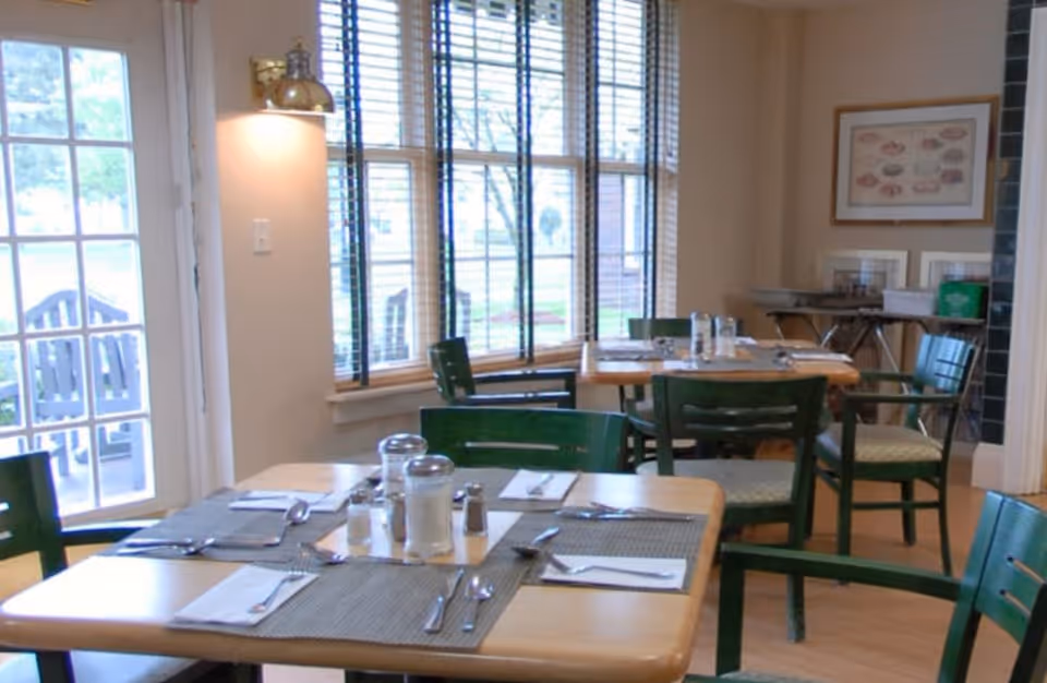 A dining area with wooden tables set with placemats, napkins, silverware, salt and pepper shakers, and glass containers. Green wooden chairs surround the tables. Large windows with blinds allow natural light to enter the room. A door with glass panes is visible on the left side, and framed artwork hangs on the wall in the background.