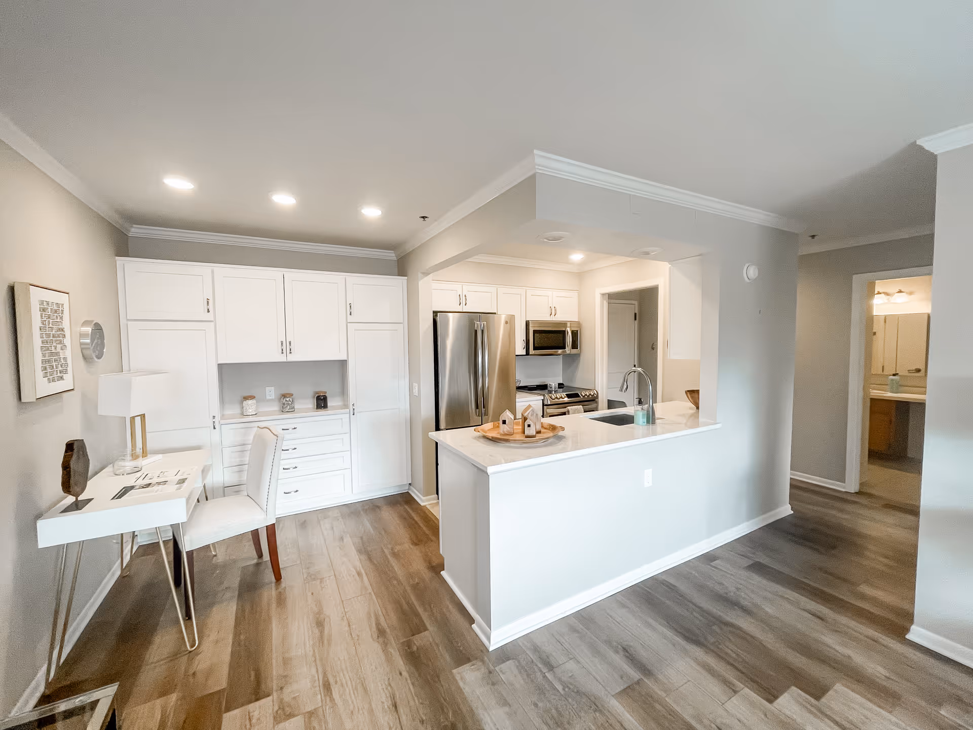 Bright and modern kitchen area with white cabinetry, stainless steel refrigerator and microwave, a white countertop island with a sink and decorative items, and a small desk with a chair and lamp against the wall. The flooring is wood-style laminate, and there is a hallway leading to another room visible in the background.