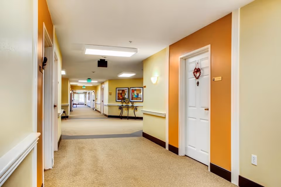 Carpeted interior hallway of a senior living facility with doors, wall art, and soft overhead lighting.