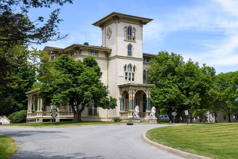 Exterior view of a large, historic-style building with cream-colored siding and ornate architectural details, surrounded by green trees and a curved driveway. Two stone lion statues flank the entrance porch.