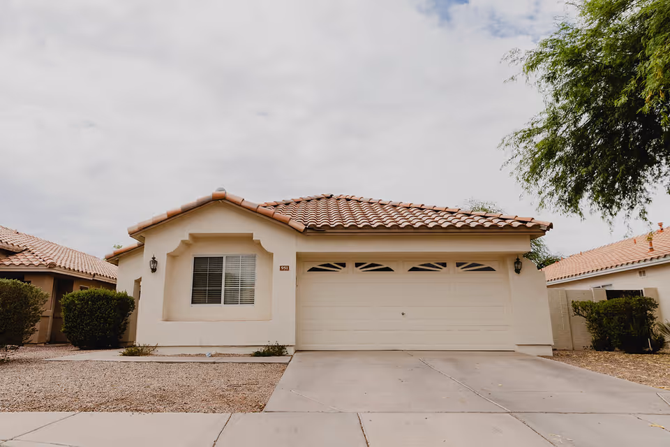 Front exterior view of a single-story house with a tiled roof, a large garage door, a window with blinds, and desert landscaping with gravel and bushes.