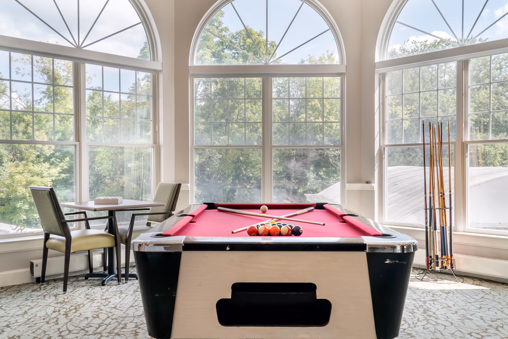 A bright room with large arched windows showing green trees outside. In the center is a pool table with a red felt surface, pool balls arranged in a triangle, and two pool cues crossed on top. To the left, there is a small table with two chairs. A rack holding additional pool cues is positioned near the right window.