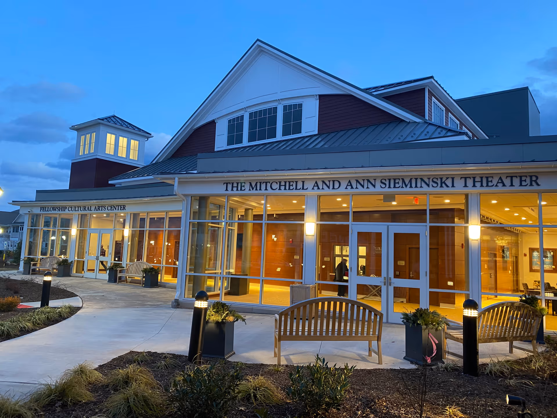 Exterior view of the Fellowship Cultural Arts Center and The Mitchell and Ann Sieminski Theater at dusk, showing a well-lit building with large windows, benches, planters, and pathway lighting.