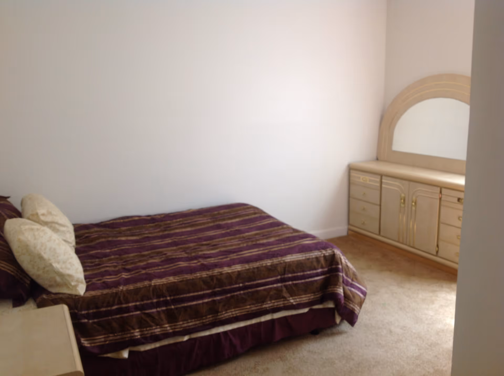 Small bedroom with a bed covered in a purple-striped comforter and a beige dresser with mirror against a white wall.