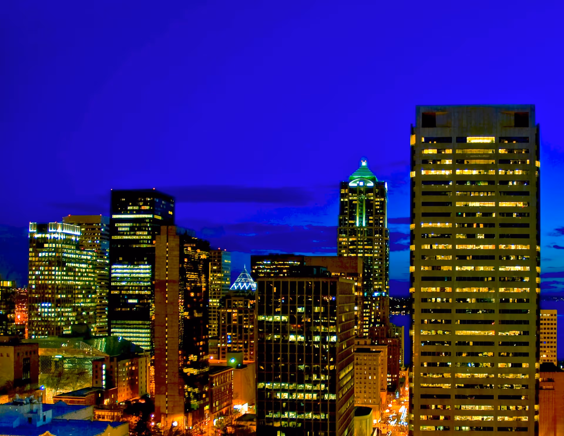 Nighttime cityscape showing illuminated skyscrapers and buildings under a deep blue sky.