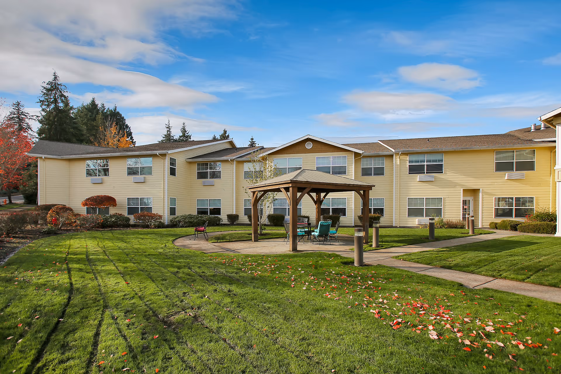 Outdoor view of Prestige Senior Living West Hills showing a two-story yellow building with multiple windows. In front of the building is a well-maintained grassy area with a wooden gazebo containing outdoor chairs and a table. The sky is partly cloudy with blue patches visible.