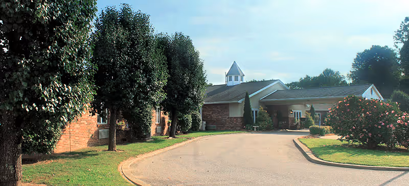 Exterior view of a single-story brick building with a peaked roof and a small cupola on top. The building is surrounded by neatly trimmed trees and bushes, with a curved driveway leading up to the entrance. The sky is partly cloudy.
