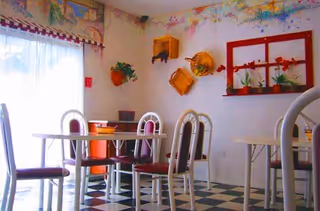 A small dining area with white tables and chairs featuring red cushions. The floor has a black and white checkered pattern. The walls are decorated with colorful floral and basket wall art, and a window with sheer white curtains lets in natural light.