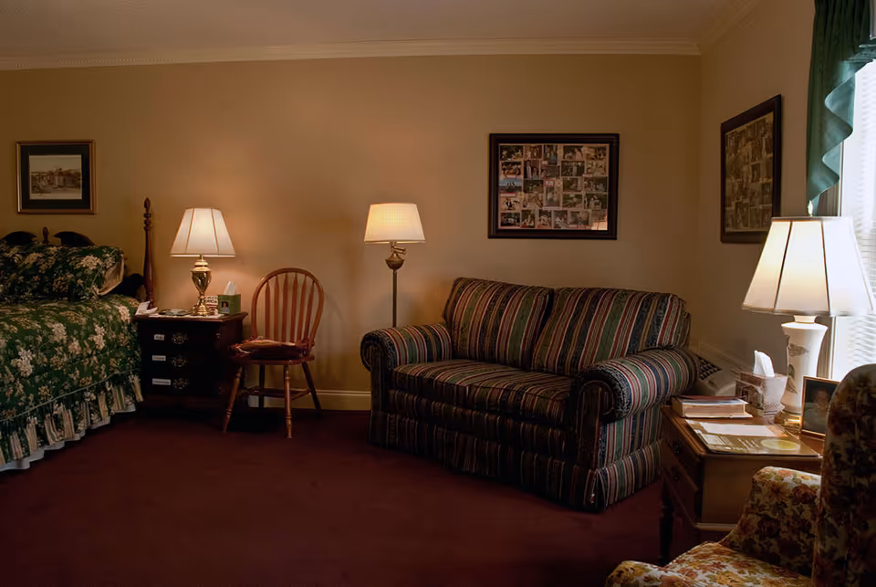 A cozy room with a floral-patterned bed on the left, a wooden chair, a striped sofa, and two table lamps providing warm lighting. There are framed pictures on the beige walls and a window with green curtains on the right side.