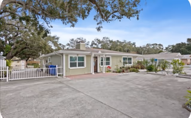 Single-story residential building with light green exterior walls, white trim, and a chimney. The building is surrounded by a concrete driveway and some landscaping with plants and trees. The sky is clear with a few clouds.