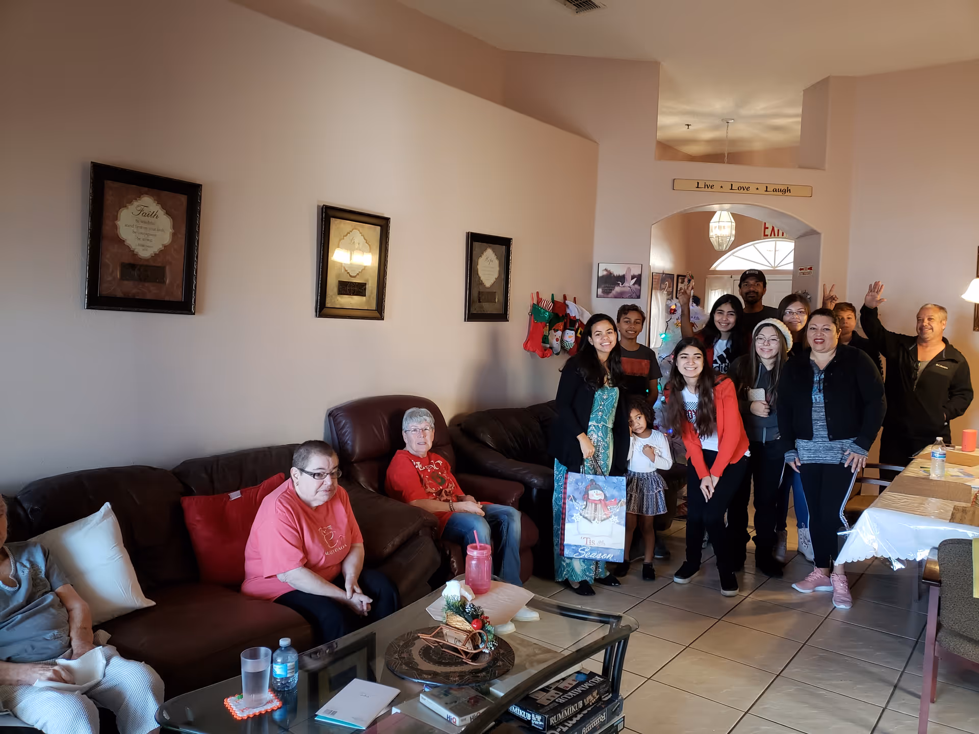 A group of residents and visitors posing in a communal living room with couches, a coffee table, holiday stockings, and a 'Live · Love · Laugh' sign.