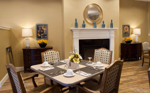 A cozy dining room with a square table set for four, featuring white cups, glasses, and silverware on placemats. The room has wooden chairs with patterned cushions, a fireplace with decorative blue vases and a round mirror above it, two side tables with lamps and yellow flower arrangements, and warm beige walls with framed sunflower artwork.