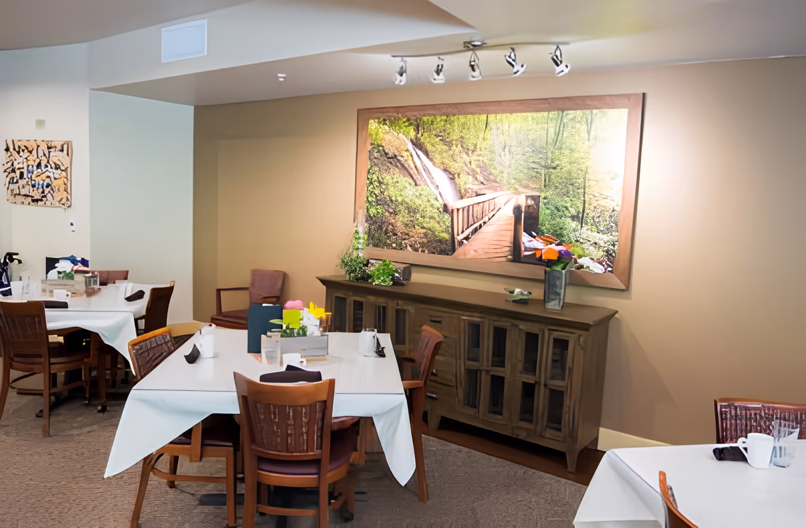 A dining room with several tables covered in white tablecloths and wooden chairs around them. On the wall is a large framed picture of a wooden bridge in a forest setting. There is a wooden cabinet beneath the picture with plants and decorative items on top. The room has beige walls and carpeted floors.