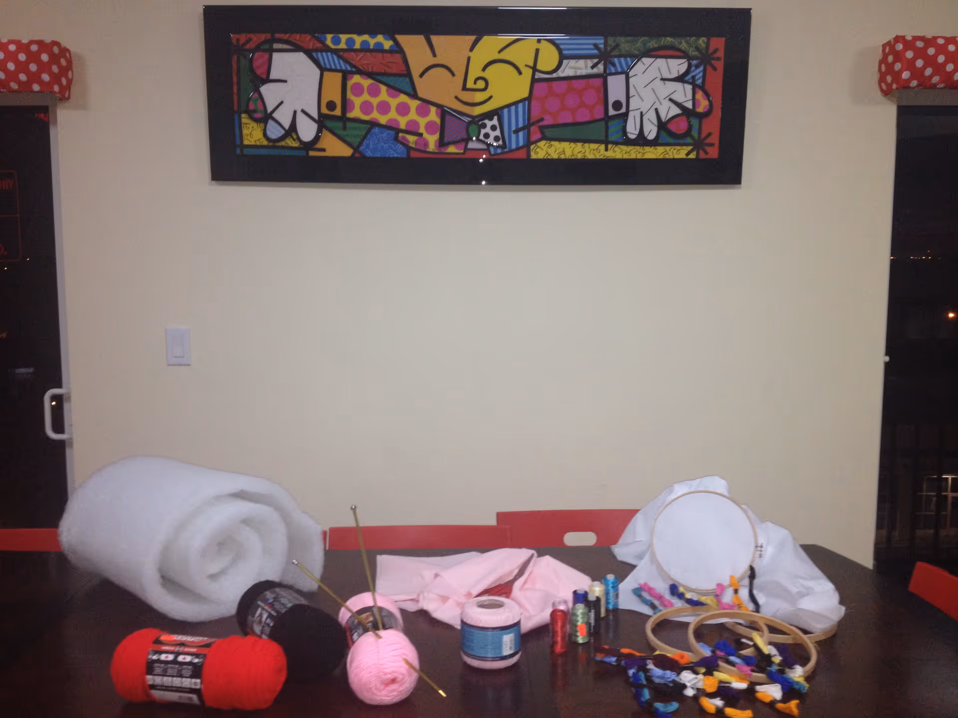 A table with various crafting supplies including yarn in red, black, and pink, knitting needles, embroidery hoops with fabric, spools of thread, and colorful embroidery floss. Behind the table is a wall with a colorful abstract painting and two windows with red and white polka dot valances.