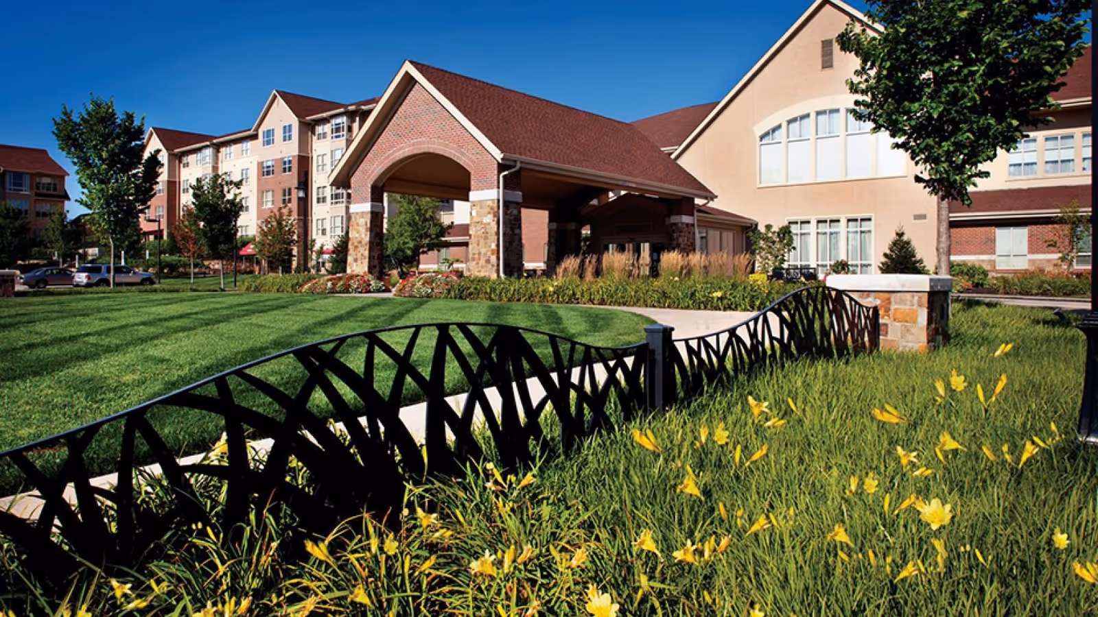 Exterior view of Tallgrass Creek Senior Living Community featuring a large building with a covered entrance, manicured green lawn, yellow flowers, trees, and a decorative black metal fence under a clear blue sky.