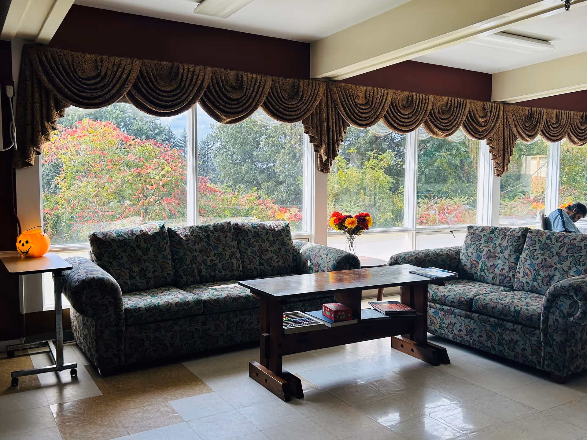 A cozy sitting area in a senior living facility with two patterned sofas facing each other and a wooden coffee table between them. Large windows behind the sofas show colorful trees outside. A small table with a pumpkin decoration is on the left, and a vase with flowers is on the right side table. A person is seated near the window on the right side.