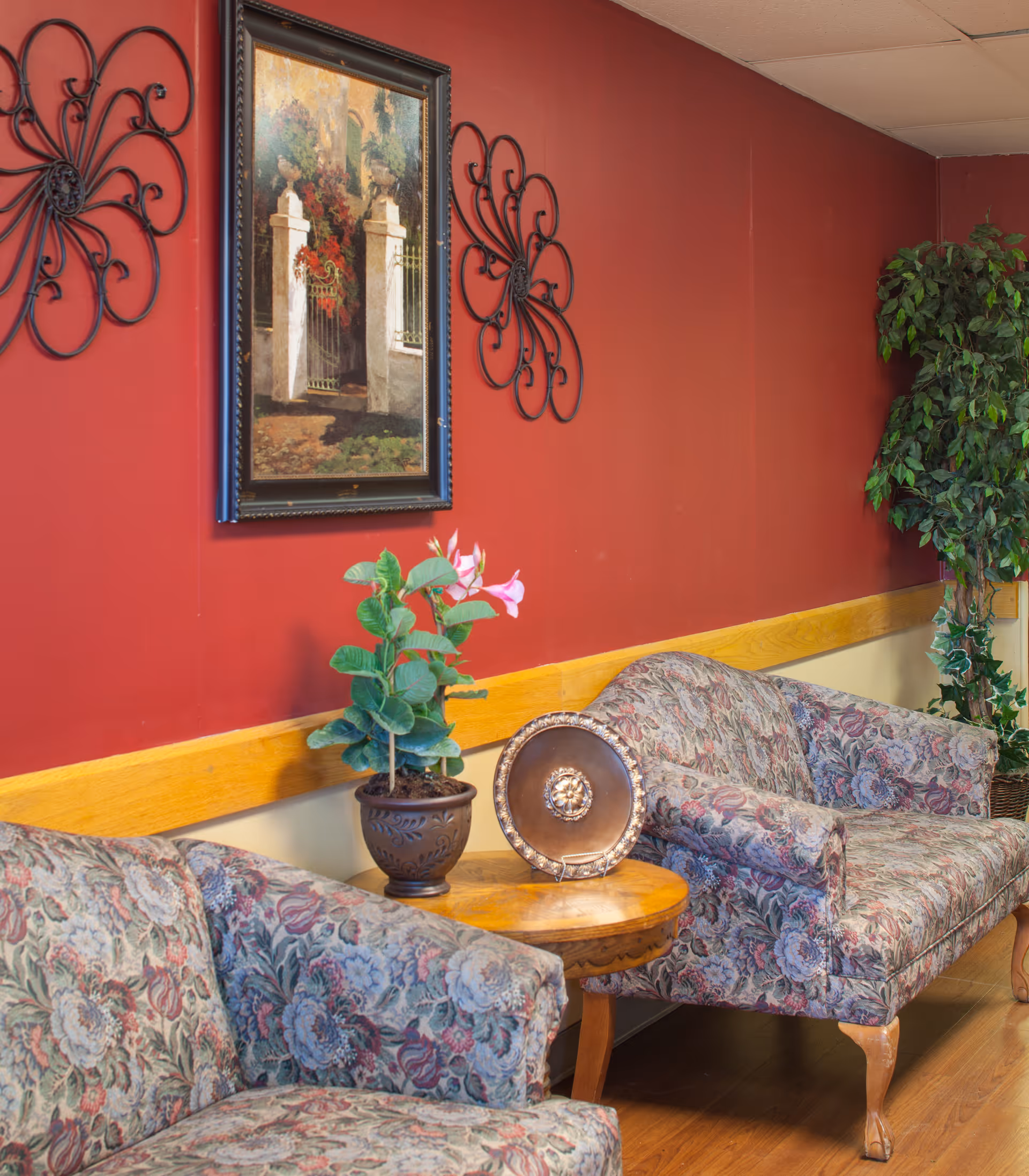 Two floral-upholstered armchairs flanking a small wooden side table with a potted plant and decorative plate against a red wall adorned with framed artwork and metal wall decor.