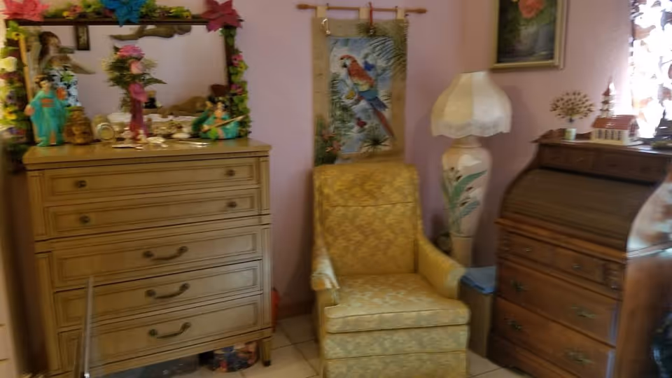 A cozy room with a yellow patterned armchair placed between a wooden dresser with a mirror decorated with flowers and a wooden roll-top desk. A tall lamp with a white lampshade and a wall hanging featuring a colorful parrot are visible on the wall behind the chair. The walls are painted light purple and there are various decorative items on the furniture.