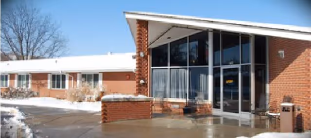 Exterior view of a single-story brick building with large glass windows and a glass door entrance. There is snow on the ground and some bare trees in the background. The building has a modern angled roof over the entrance.