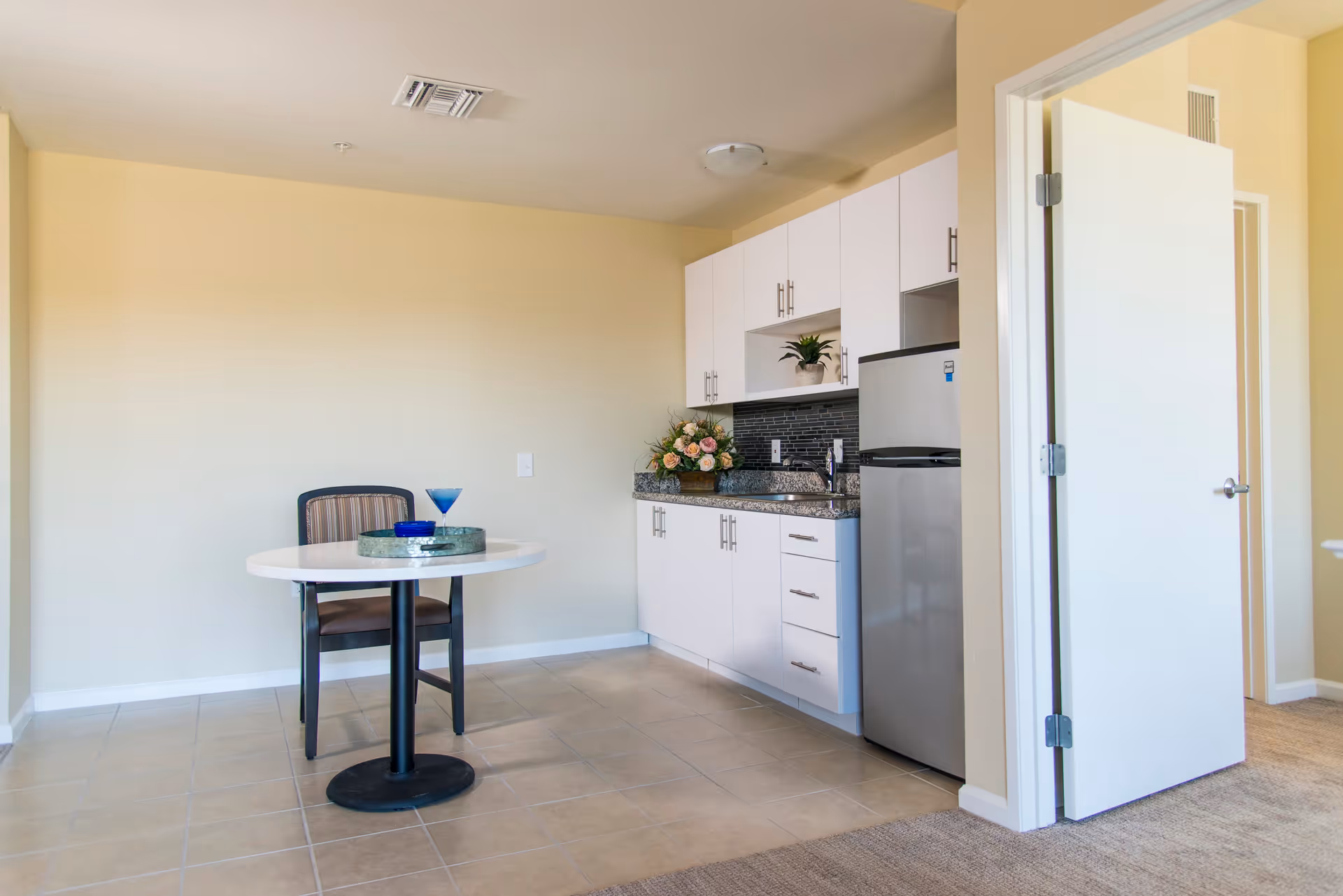 Small kitchen area with white cabinets, a granite countertop, a stainless steel mini refrigerator, and a sink. A round table with a single chair is positioned nearby, with a blue glass and a decorative tray on the table. The room has beige walls and tiled flooring, with an open door leading to another carpeted room.