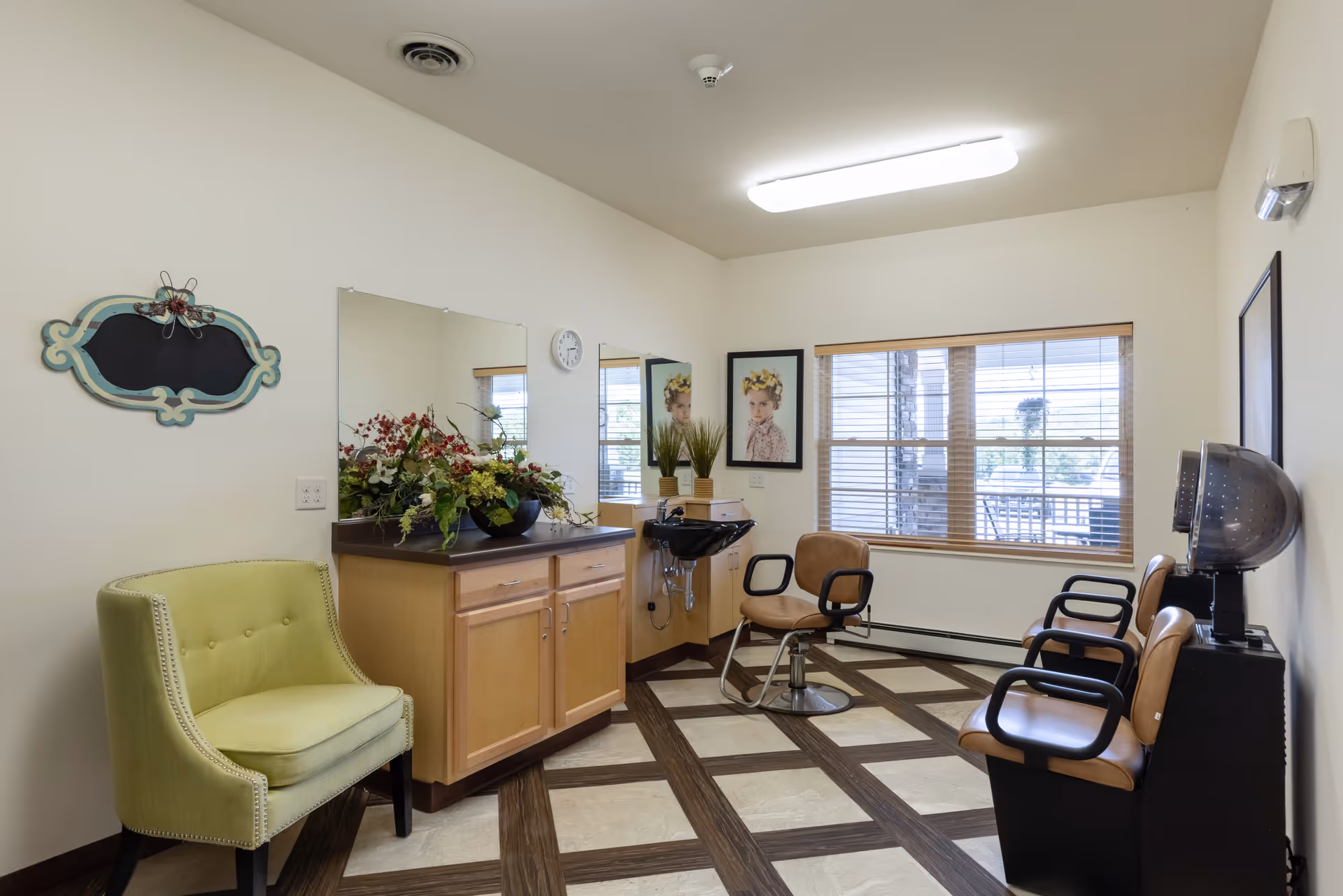 Interior of a small salon room with a green upholstered chair, a wooden cabinet with a large mirror above it, a black hair washing sink, three salon chairs, a hair dryer, and a window with blinds letting in natural light. The room has light-colored walls and a patterned floor.