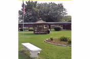 Outdoor area of a senior living facility featuring a green lawn, a white stone bench, a garden bed with plants, an American flag on a pole, and a wooden gazebo with seating under it. The background shows a single-story building with a covered walkway and trees.