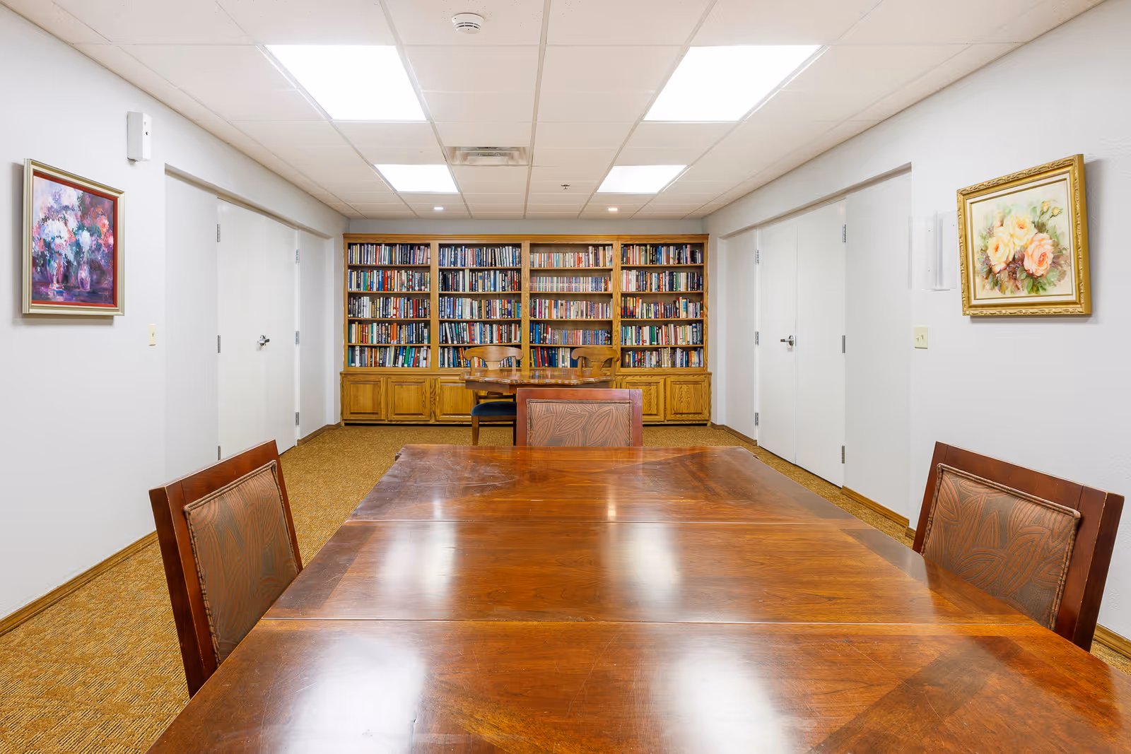 A well-lit room with a large wooden table and chairs in the foreground. At the back of the room, there is a wooden bookshelf filled with books. The walls are white and decorated with framed floral paintings. The floor is carpeted in a light brown color.
