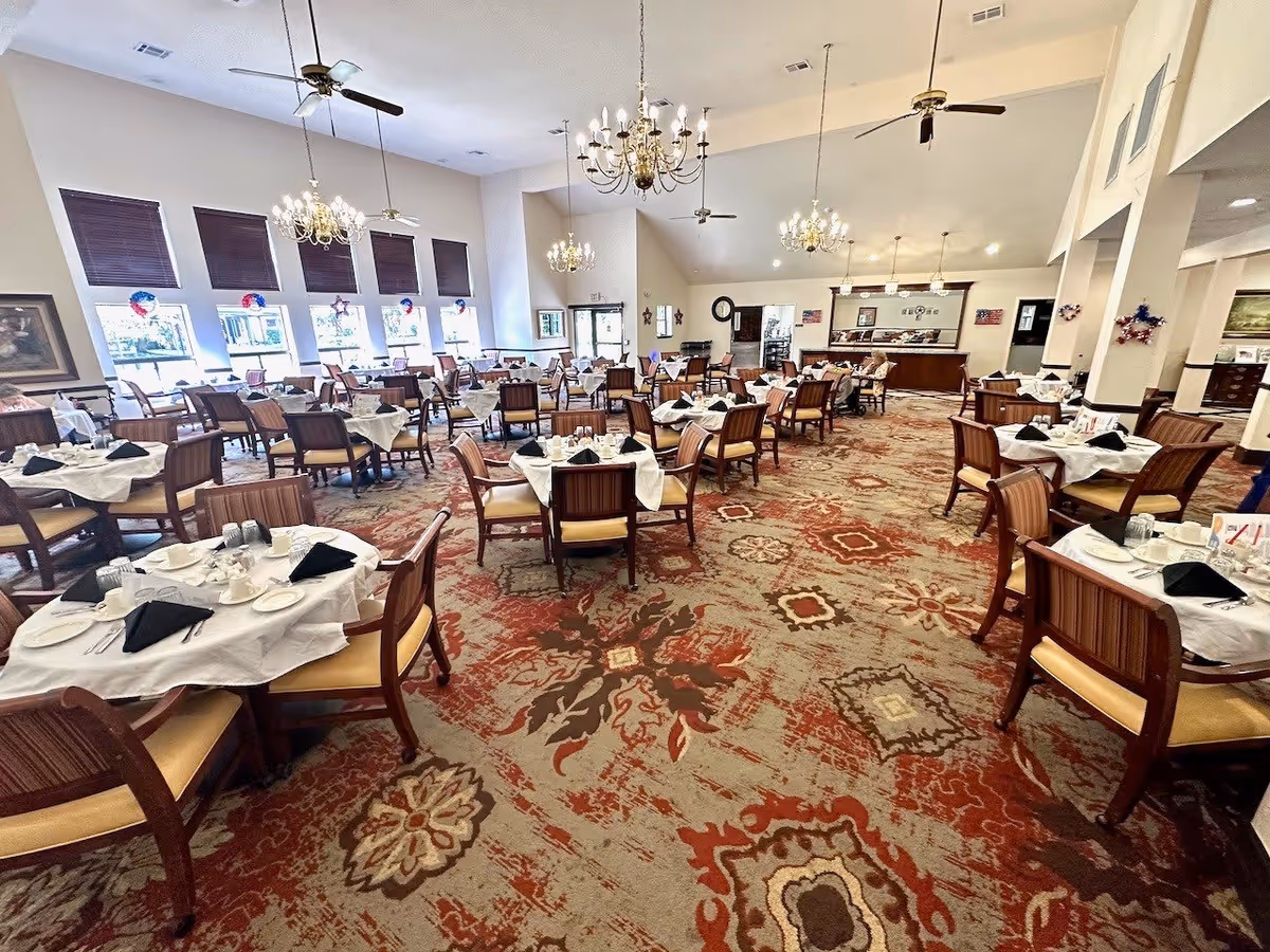 A spacious dining room with multiple round tables covered with white tablecloths, each set with plates, cups, utensils, and black napkins. The room features large windows with dark blinds, ornate chandeliers hanging from a high ceiling, ceiling fans, and a patterned carpet with red, brown, and beige tones. There is a counter area at the back of the room with a mirror and some decorations on the walls.