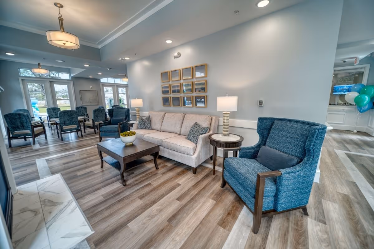 A bright and spacious living room area in an assisted living facility featuring a beige sofa with two patterned pillows, a blue upholstered armchair, a wooden coffee table with a decorative bowl, and a side table with a lamp. The room has wood-look flooring, large windows letting in natural light, and framed artwork on the wall. Additional seating and tables are visible in the background.