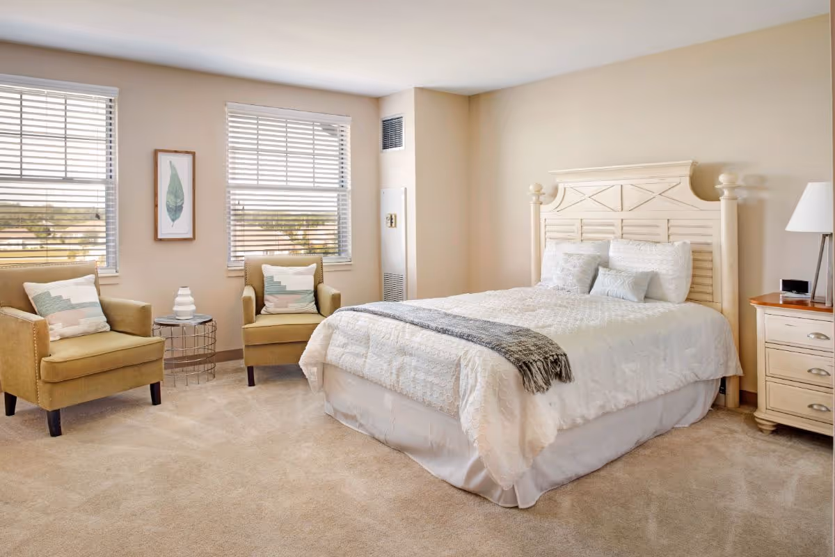 A bright bedroom with a large bed featuring white bedding and multiple pillows. Two beige armchairs with patterned cushions are positioned near two windows with blinds. A small round side table with a decorative vase is placed between the chairs. A wooden nightstand with a lamp and an alarm clock is next to the bed. The room has beige walls and carpeted flooring.