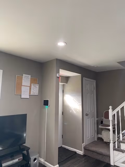 Interior view of a room with gray walls, a flat-screen TV on a stand, a bulletin board with papers on the wall, a doorway leading to another room, and a stairlift installed on a staircase.