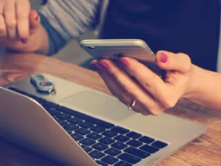 Close-up of hands using a smartphone over a laptop on a wooden desk.